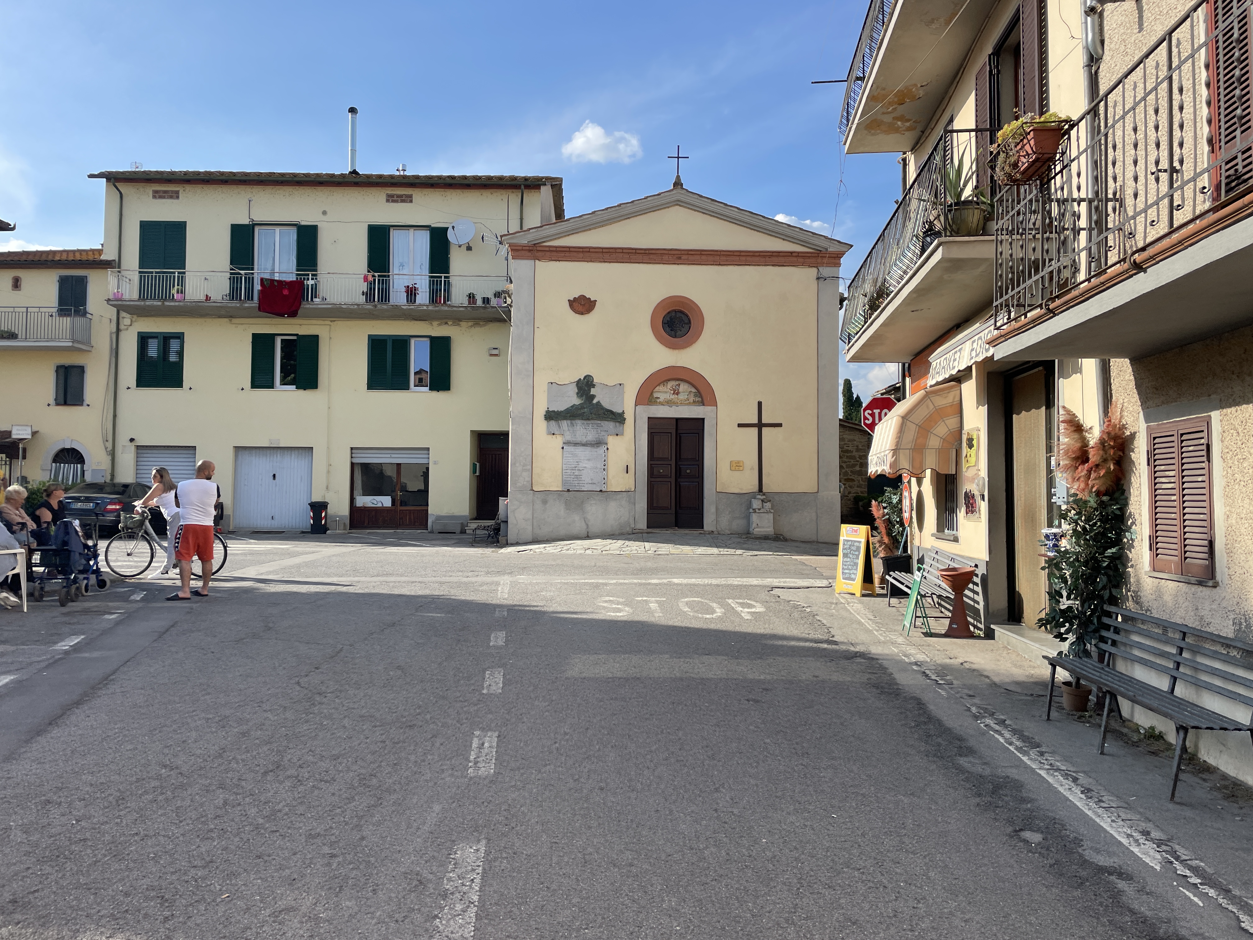 Road leading into Borghetto Square. Beige church with red portal and wooden cross on right side. On the left, some houses.